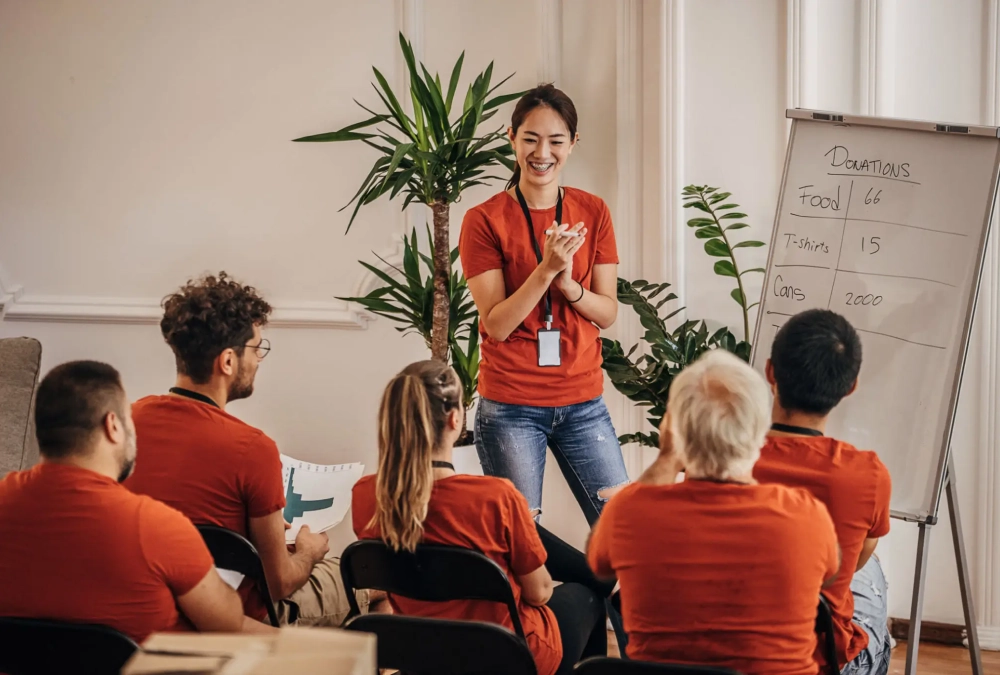 A woman enthusiastically speaking to a seated group in a bright room.
