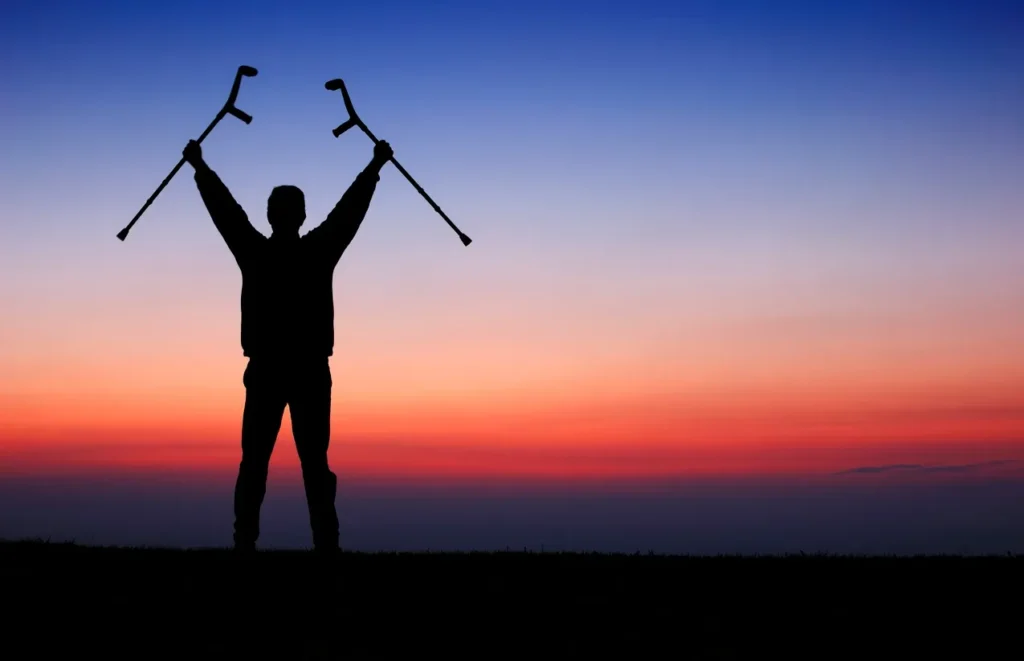 Silhouette holding crutches against sunset sky.