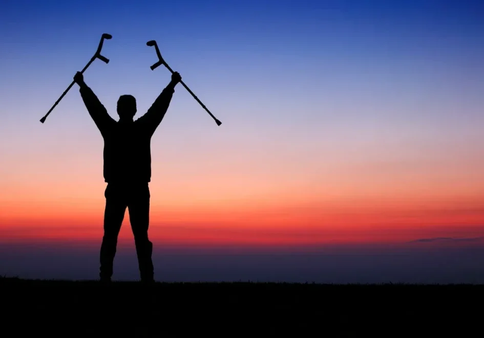 Silhouette holding crutches against sunset sky.
